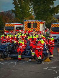 Gruppenfoto von Azubis zum Notfallsanitäter (m/w/d) bei einer Übung: Azubis sitzen auf einem Wagen, bei dem das Dach fehlt, und stehen um den PKW herum. Im Hintergrund stehen Rettungsdienstfahrzeuge.